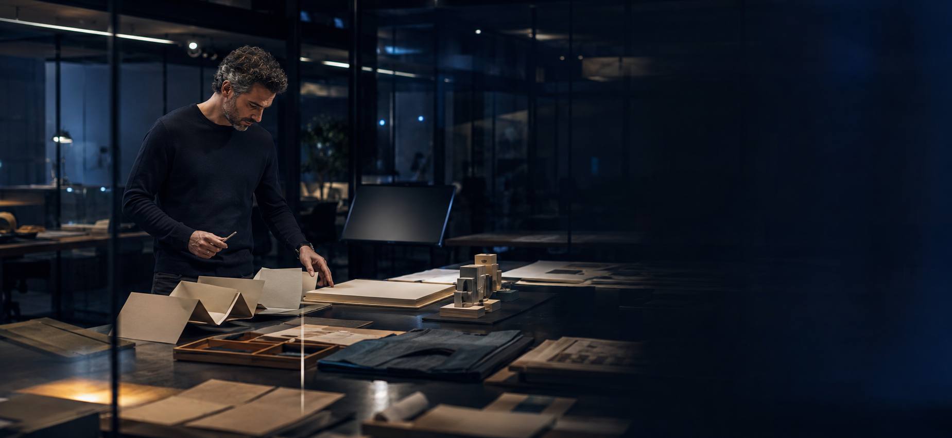 A creator standing at a materials table surrounded by papers, samples and an ambiguous prototype, deep negative space to the right